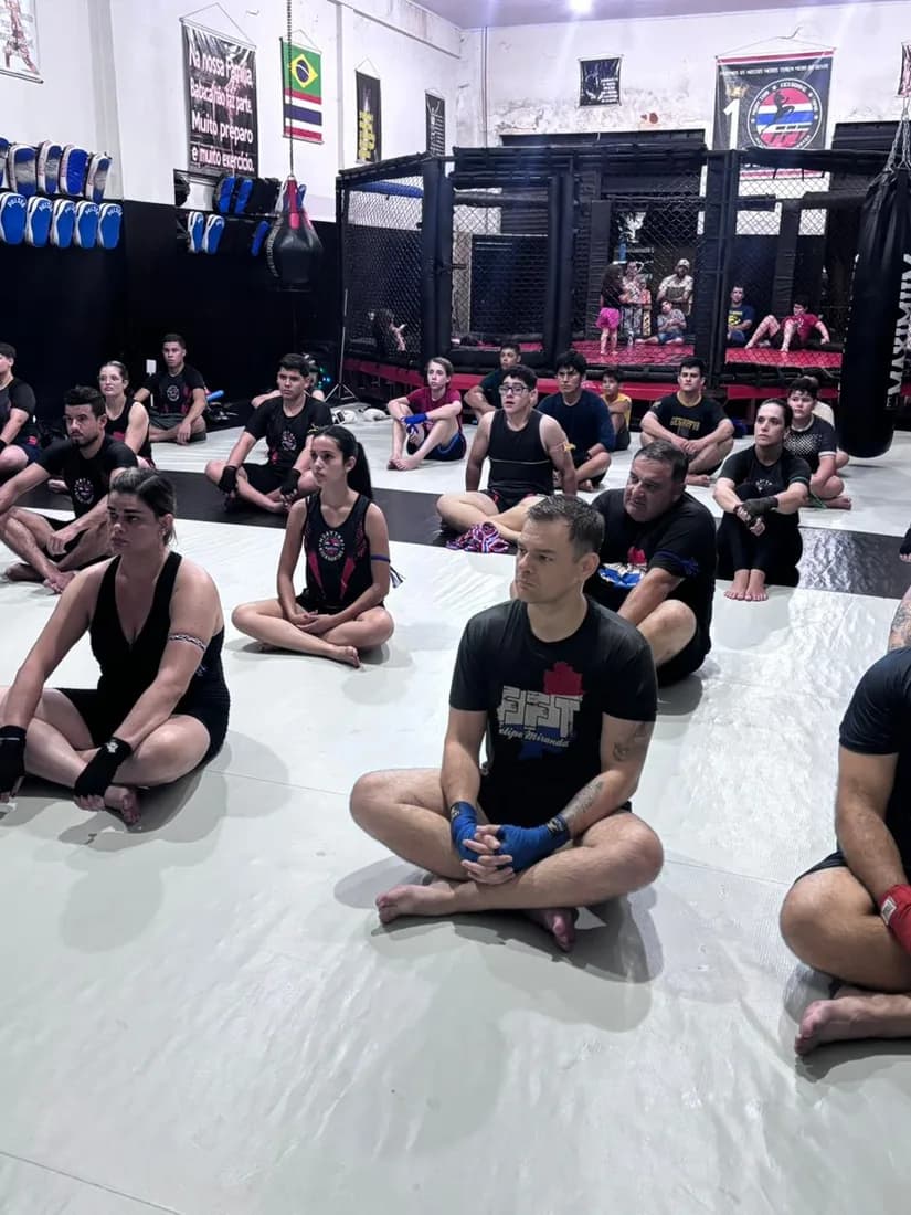 Alunos de várias idades sentados no tatame branco da academia da Equipe FFT durante a aula