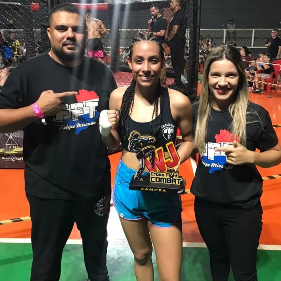 Atleta da Equipe FFT segurando o cinturão de campeã ao lado da equipe técnica dentro da arena